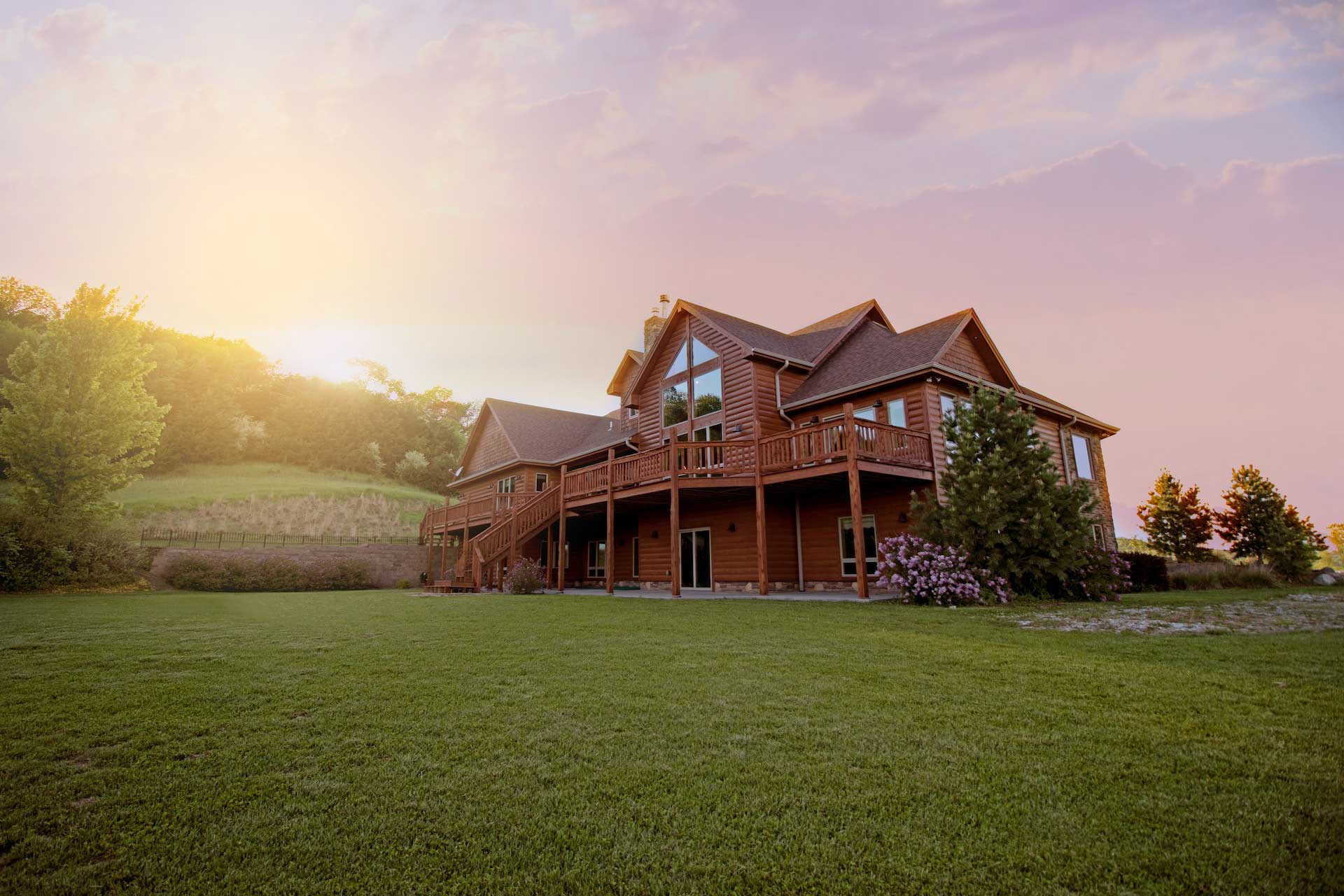 A log home with wrap around porch seen from across a green lawn with sunlight streaming in from the left